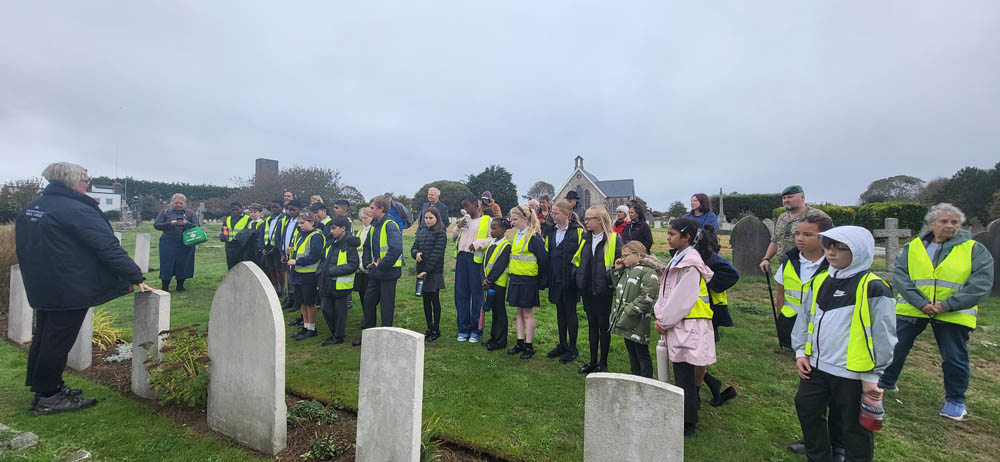 Schoolchildren listen to a talk given by a CWGC public engagement coordinator in Seaford Cemetery, Essex, in front of a row of headstones.