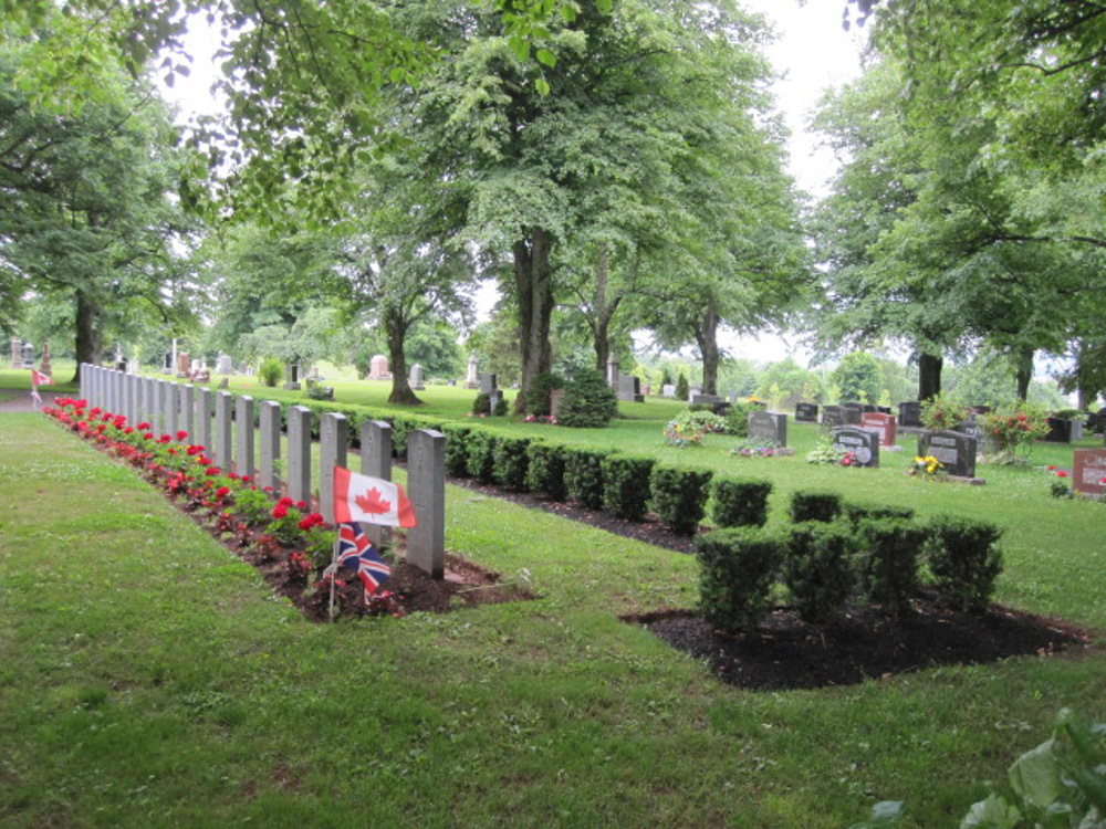 Row of CWGC headstones sits in front of a decorative hedge. The plot of each headstone features a vivid red rose. A Canadian flag above a union jack has been placed at the end of the headstone row.