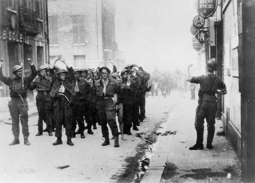 A German soldier directs a group of Canadian prisoners of war through the streets of Dieppe circa August 1942. The Canadians are marching in a line three-men across and about six deep with their hands raised above their heads in surrender. Some of the Canadian soldiers wear bandages after being wounded in previous combat.