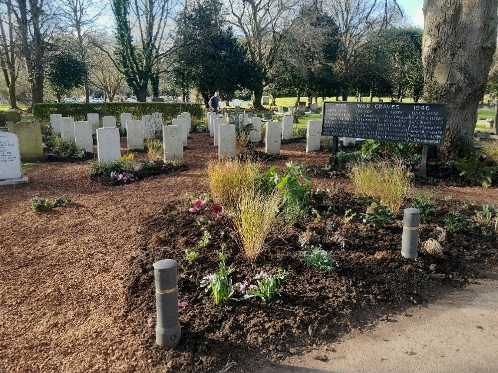 CWGC headstones sitting on a woodchipped plot. In the foreground are bright plants sat in a turf planter. A big black sign with war grave information in white lettering is visible resting beneath a tree.