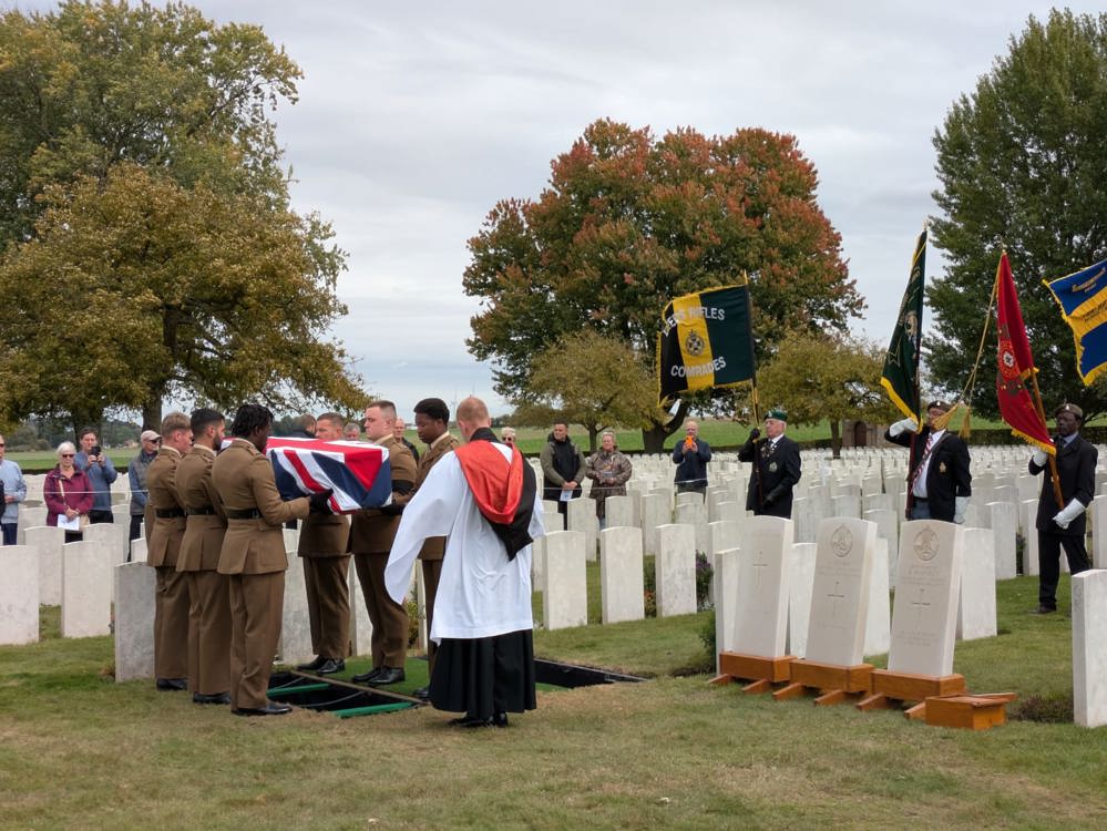 British soldiers lower a union-jack shaped coffin into a war grave in Warlencourt British Cemetery. A series of CWGC headstones waiting installation on wooden posts are visible to the right. In the background, standard bearers hold various military banners.