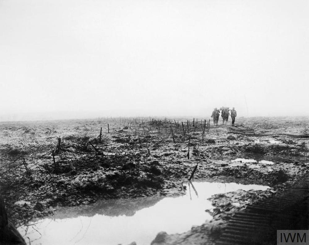 Canadian stretcher bearers carry a wounded soldier on their shoulders over a shell-torn landscape. The ground is thick and muddy, with deep shell craters full of muddy rain water visible in the foreground.