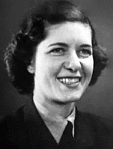 Section Officer Yolande Beekman. She is smiling a happy grin and has wavy dark hair that falls in tuffs around her ears. She is wearing a dark uniform with a light shirt.