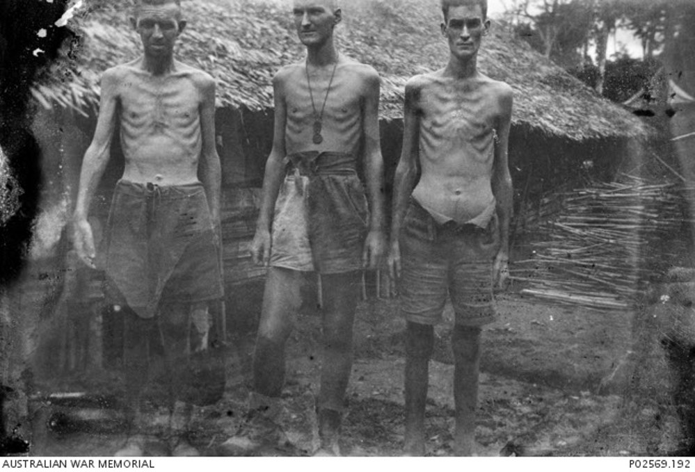 Three emaciated Australian Japanese POWs pose in front of low reed and thatch hut. Their ribs and skeletal structure are clearly visible, the results of the terrible treatment at the hands of the Imperial Japanese.