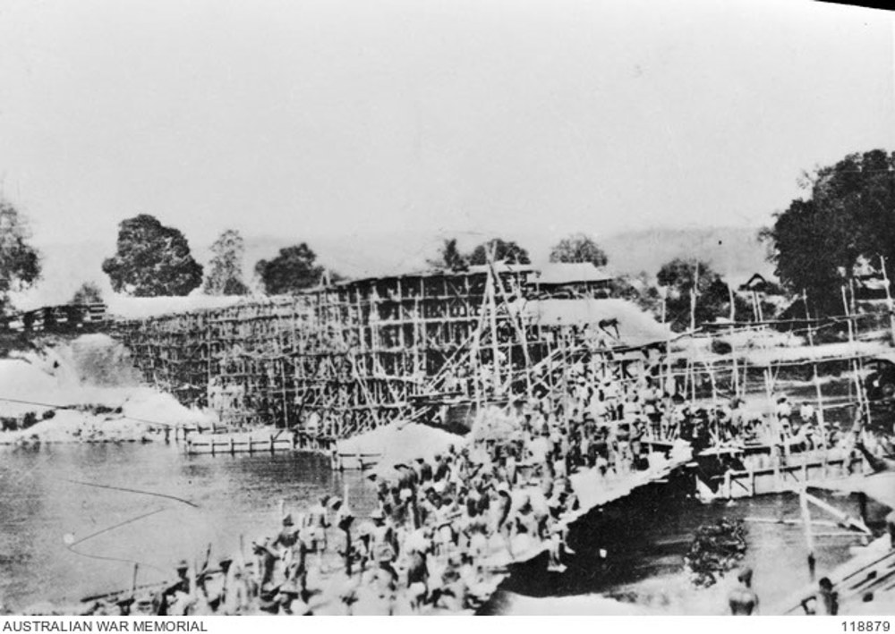 Hundreds of Allied prisoners of war constructing a railway bridge over a river in Thailand during the Second World War. The bridge is built of bamboo and wood slats and poles, looking more like scaffolding thant a completed bridge. The river coursing beneath is wide. Trees flank both banks.