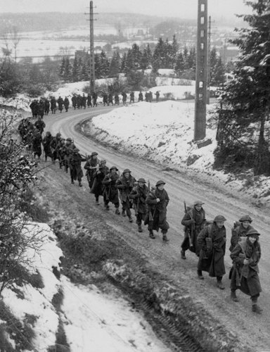 A column of British WW2 era soldiers moves up a curving road in the Ardennes during the Battle of the Bulge. The road has been cleared by the banks on either side are covered in snow. Telephone pylons and snow-covered trees flank the road.