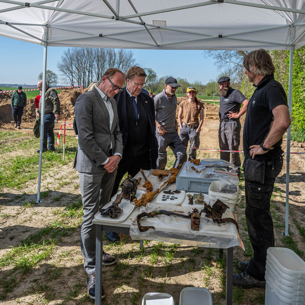 Suited men examine a table of rusted and weathered First World War artefacts extracted from an archaeological dig site in Belgium. The table as been set up under a wide white tent. Workers and a large dirt pile can be seen in the background.