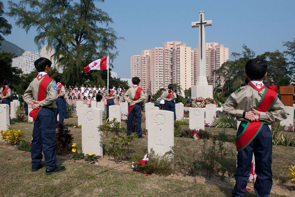 Schoolchindred in green-tipped red sashes and khaki and navy blue school uniforms stand to attention next to the cross of sacrifice at Sai Wan War Cemetery Hong Kong for Remembrance Day.