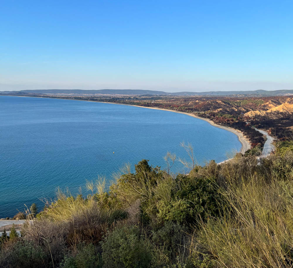 A low cove seen from scrubland in Gallipoli. A sandy beach circuits the deep blue sea. Mountains are visible in the background as the beach gives away to rugged scrubland.