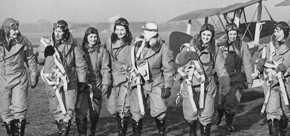 A row of female aviators walking away from a bi plane. They are wearing flight suits with leather goggles and hats.