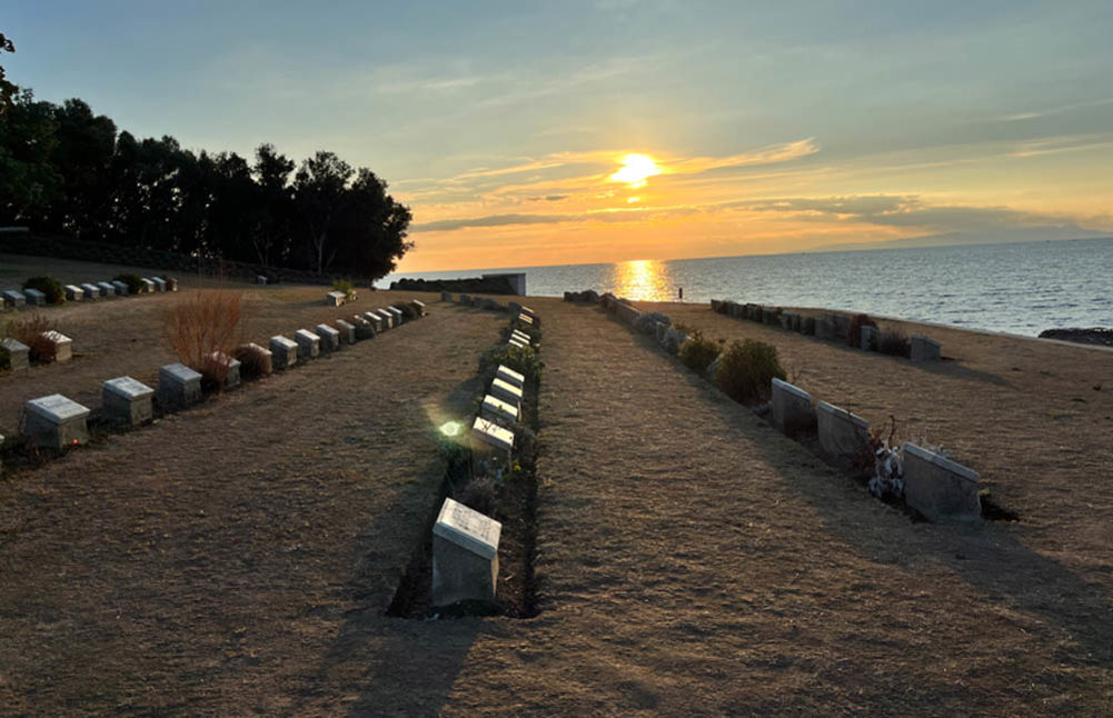 The sun setting on a low row of headstones at Ari Burnu Cemetery, ANZAC. The sun breathes war orangey hues into the blue sky with the orb of light reflecting off the sparkling waves of the slate blue Aegean.