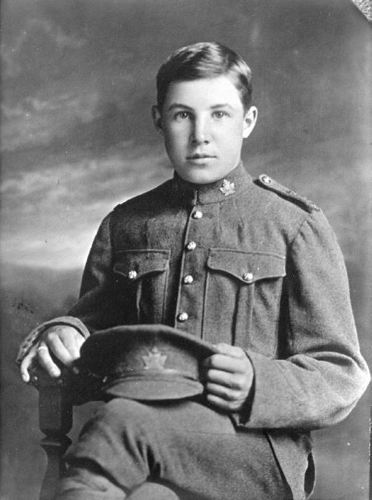 Private Clifford Oulton sits on a wooden chair posing for a photograph. He is very young, aged just 14. Maple leaf badges are visible on his uniform's collar. He is holding a peaked cap with a maple leaf badge on his lap.