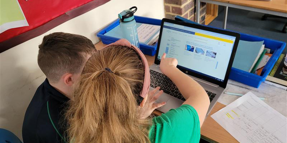 Over the sholder view of a teacher in a green shirt showing a male pupil how to navigate the CWGC website on a laptop.