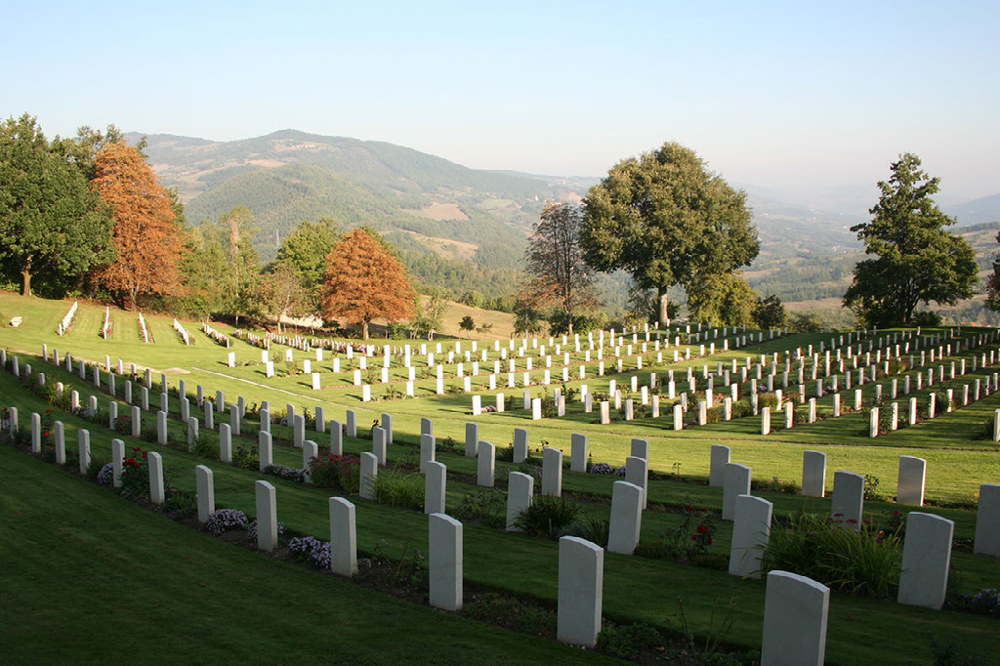 Castiglione War Cemetery showing rows of CWGC following a gentle slope towards tall trees in autumnal orange and evergreen. Mountains are visible in the background.