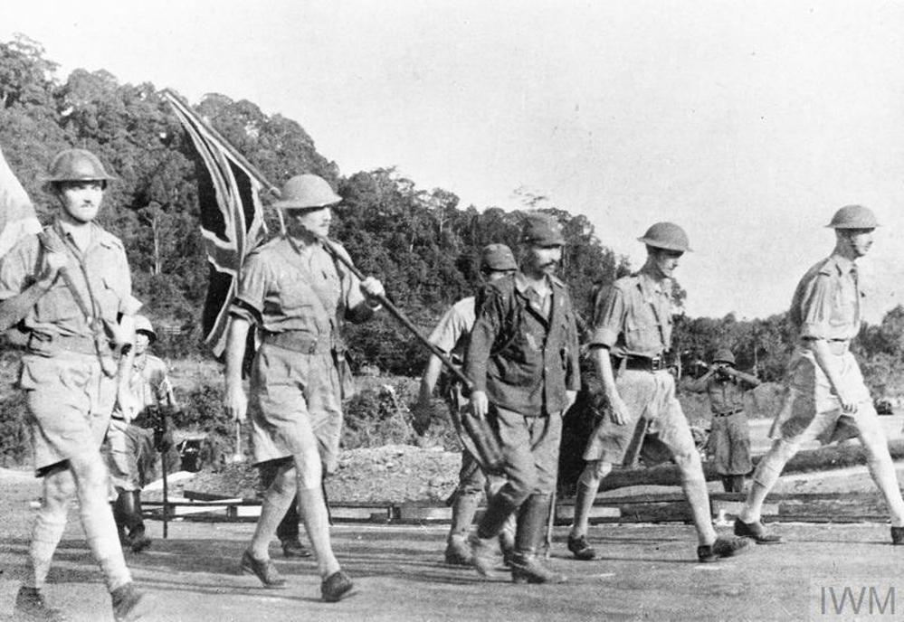 British soldiers, two carrying union jack flags via poles resting on their shoulders, are escorted by Japanese soldiers on their way to surrender in Singapore, early 1942.