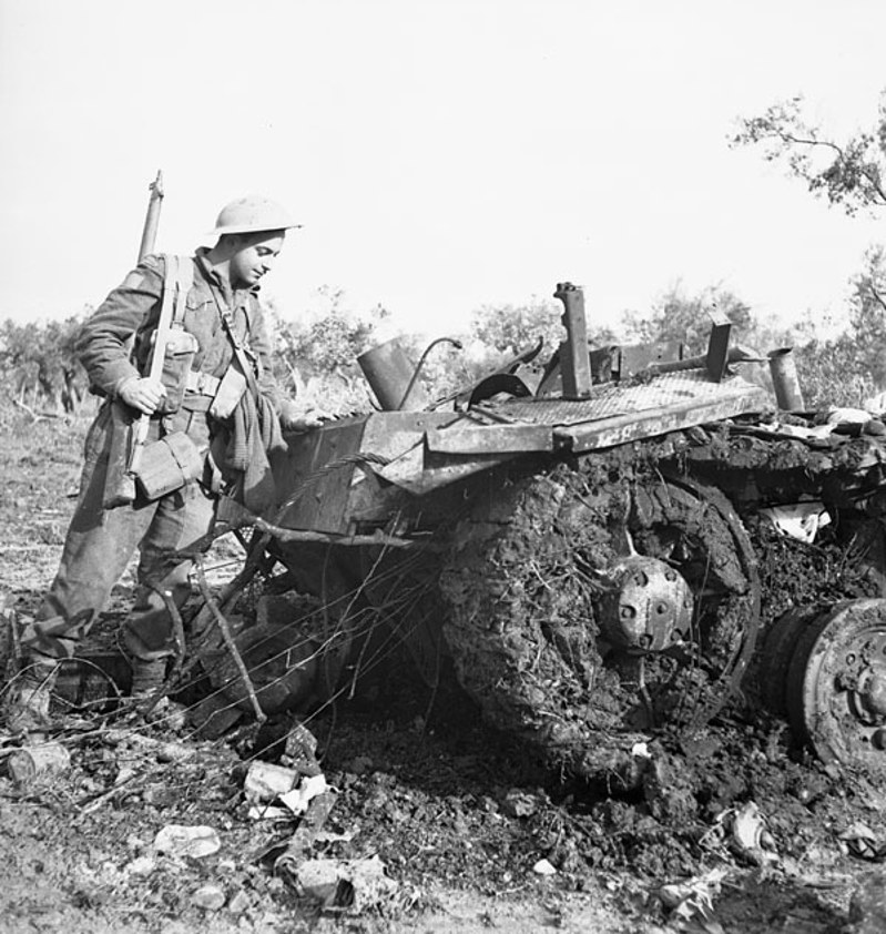Canadian WW2 solider holding a rifle stands next to the smashed and twisted wreckage of a German vehicle.