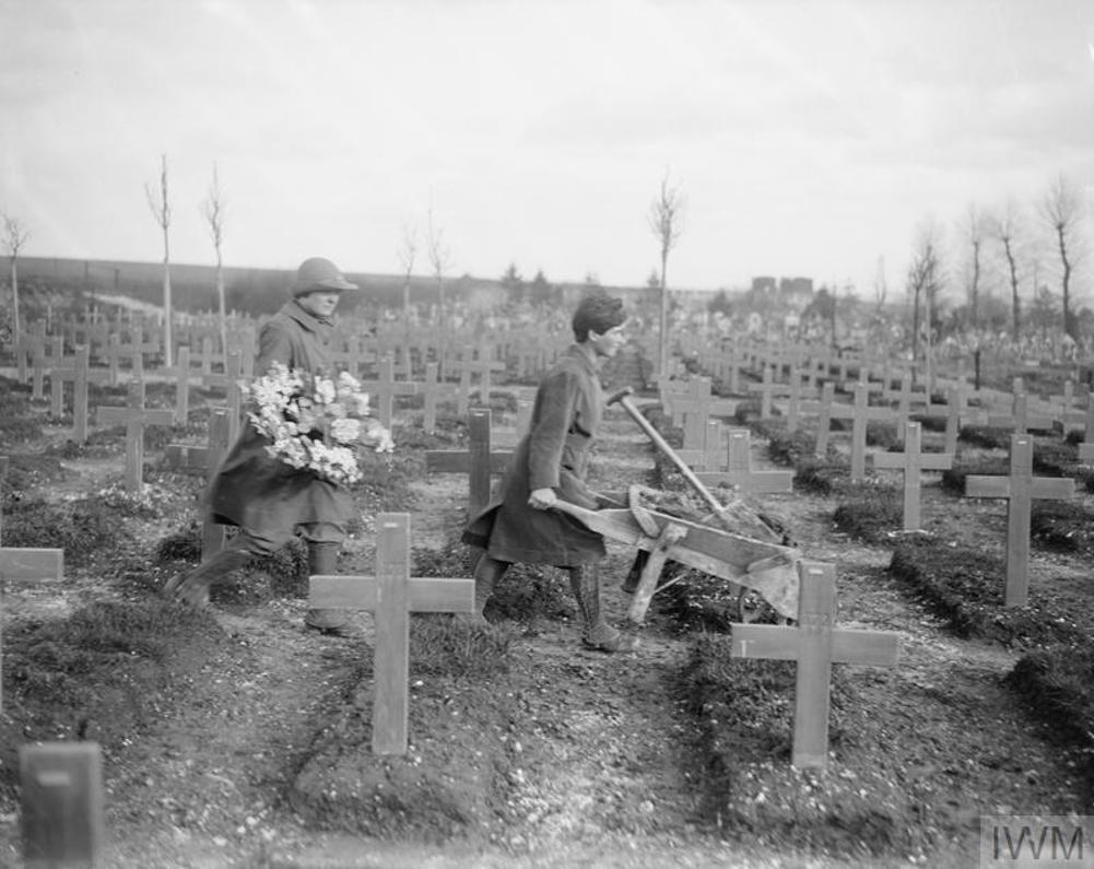 Two members of the Women's Auxiliary Army Services tend to graves in Abbeville Cemetery, France, in February 1918. Both women are wearing military great coats and helmets. The first women pushes a wheelbarrow while the second follows behind carrying a bouquet of flowers. They are walking through a cemetery with war graves marked with wooden cross markers. The rows stretch on into the background. The ground is still relatively freshly dug or churned in places.