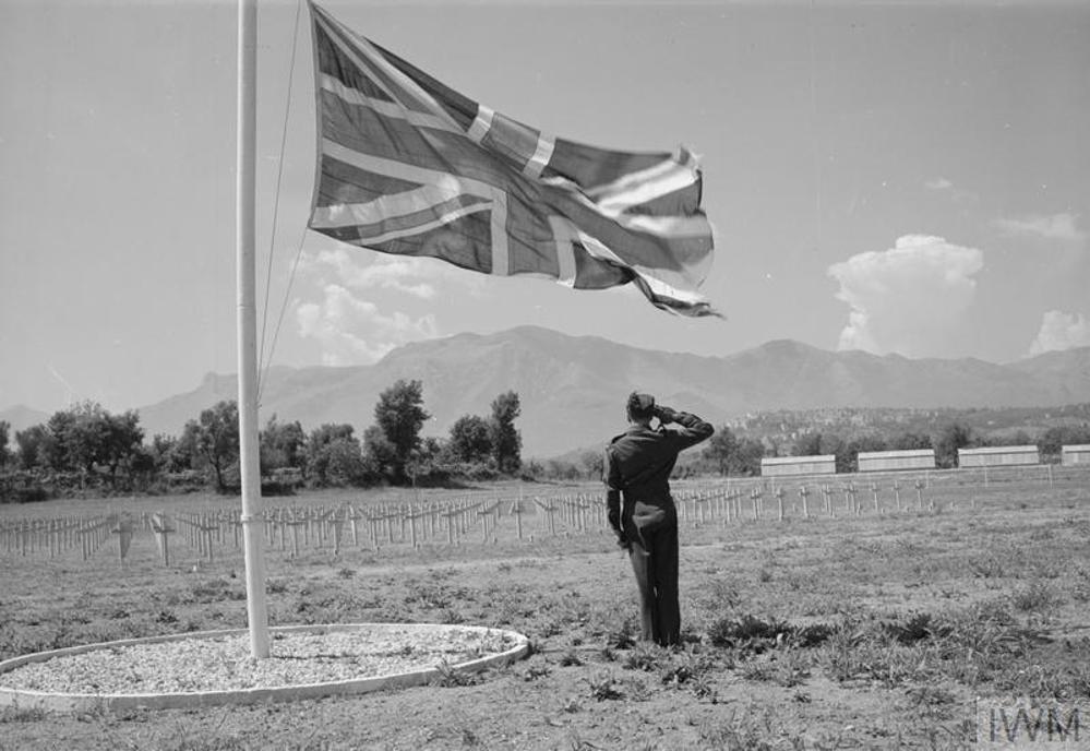 A solo British serviceman salutes the war graves at Minturno War Cemetery. He stands beneath a large union jack flag hung from a white flagpole. The war graves are marked by rows of white wooden cross-shaped grave markers.