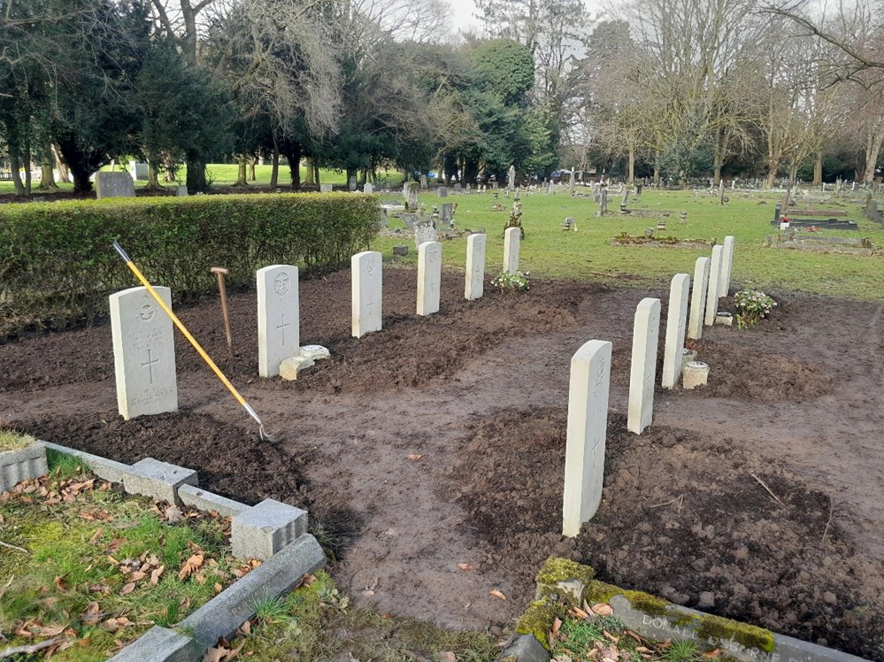 A CWGC headstone plot. Each headstone row geatures six white limestone headstones with carved crosses and regimental badges. The turf has been dug up ready for planting. A hoe rests against the back rest headstone.