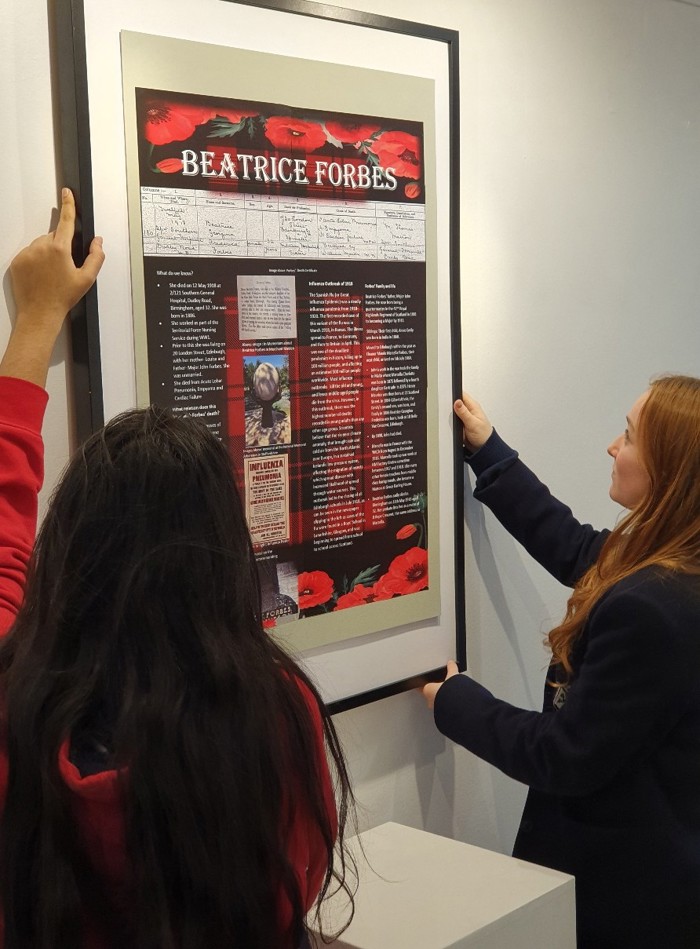 Two girls mount an exhibition poster on a wall. The words BEATRICE FORBES are visible on the poster as are several columns of text set over a black and red poppy background.