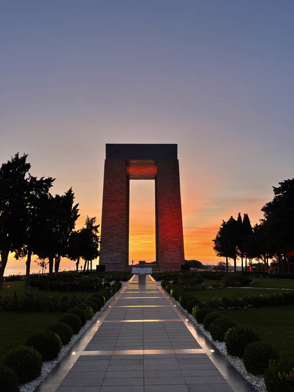 The Turkish Martyrs Memorial Gallipoli set against a setting sun. A warm orange glow illuminates the twin-pillared memorial, blending into the low lilac and blue sky. Two rows of trees flank a stone pathway leading up to the memorial.