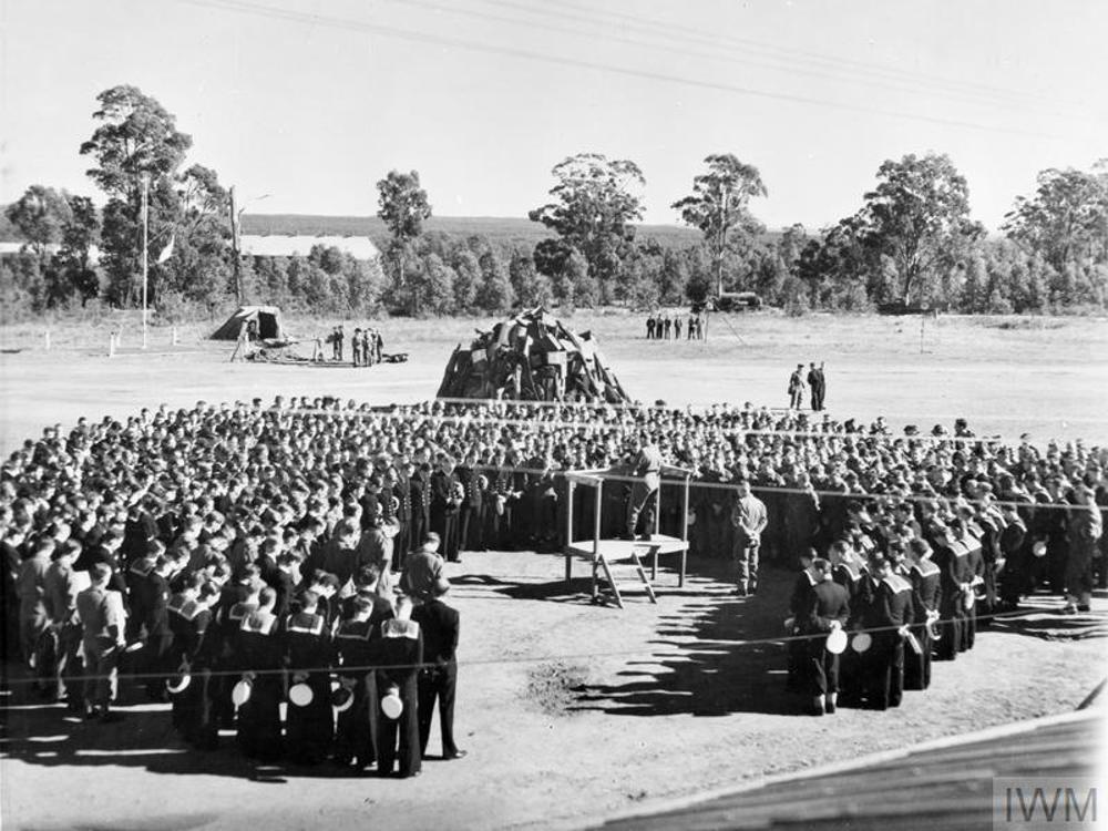 Australian soldiers and sailors attending a sombre commemorative ceremony. The trroops are gathered around a wooden rostrum in a semi circle. On the rostrum, a military padre gives a sermon.