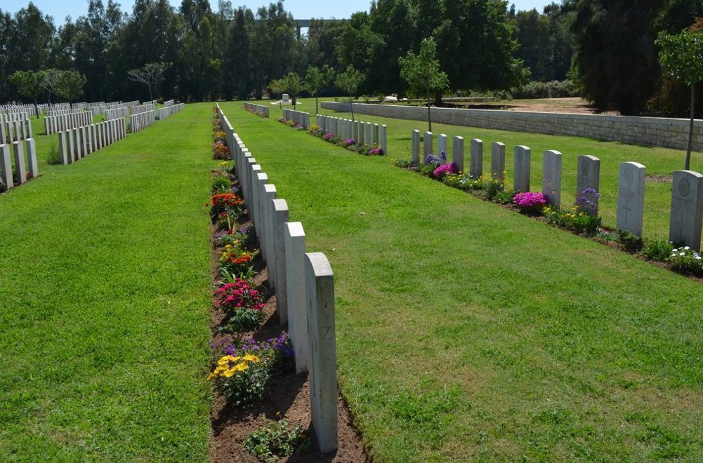 Headstone rows featuring blooming plant borders and a lush green lawn at Ramleh War Cemetery.