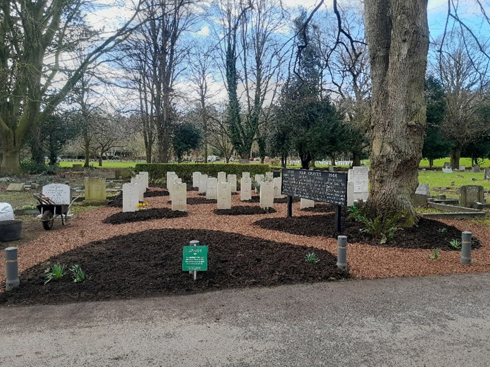 A plot of CWGC headstones sits on a mixture of turf and gravelled paths. Fresh turf for planting is visible in front of some of the headstones, as well as the foreground planter, and underneath a large tree. To the left is a wheelbarrow filled with gardening tools.