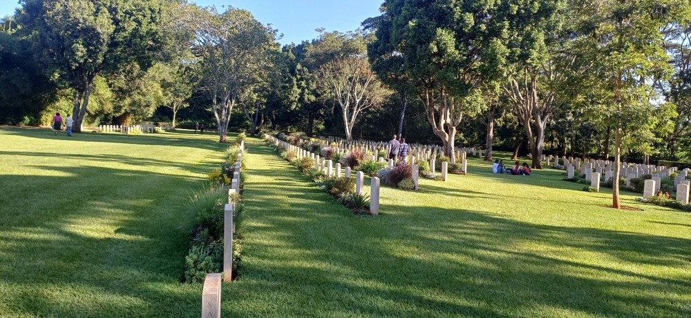 Rows of headstones underneath various species of tree on a lush green lawn in Nairobi War Cemetery.
