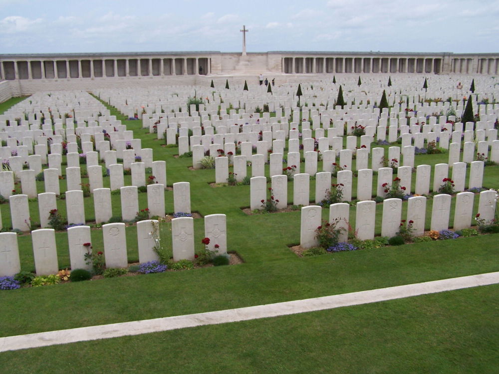 Pozieres British Cemetery showing rows of Portland stone CWGC headstones leading up to a colonaded memorial at the rear of the site. The Cross of Sacrifice sits in a gap in the memorial wall in the centre of the rear wall.