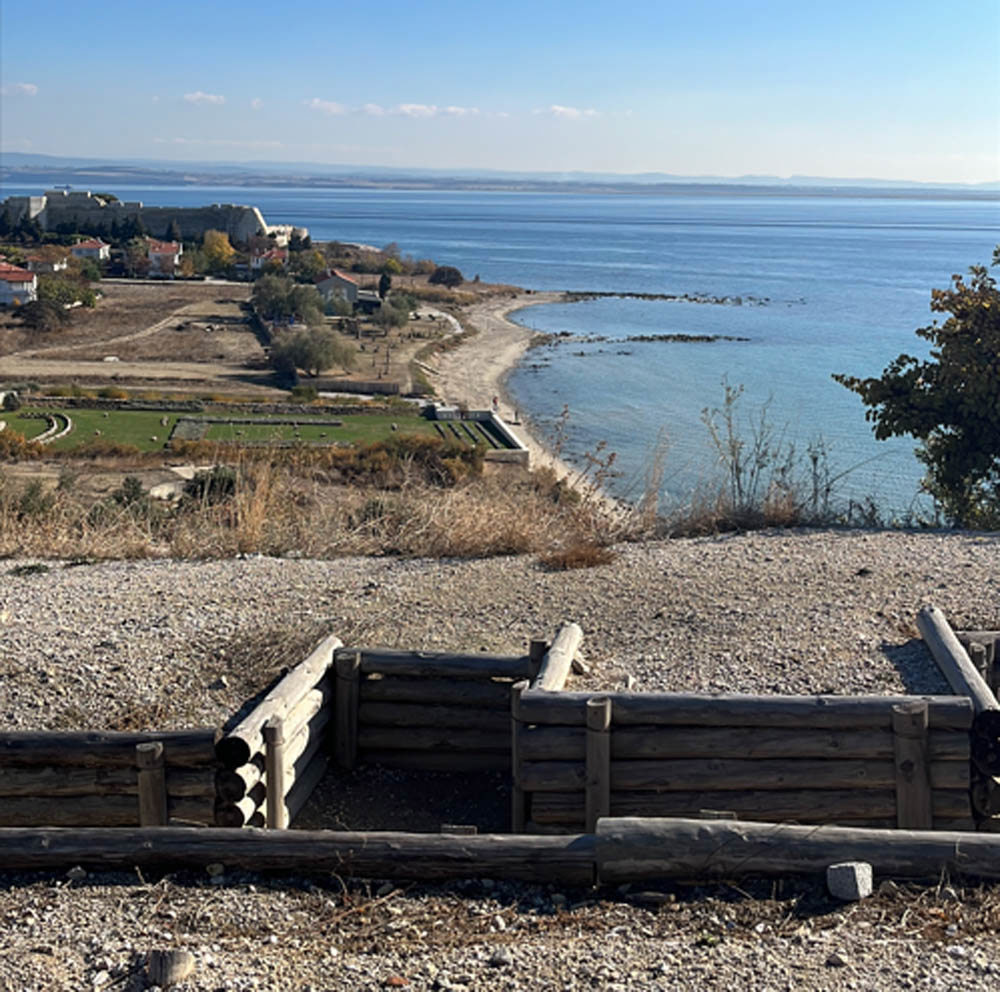 Remains of a Turkish trench network overlooking the Aegean Sea on the Gallipoli Peninsula. A verdant green war cemetery, studded with rows of white headstones, is visible from the trench.