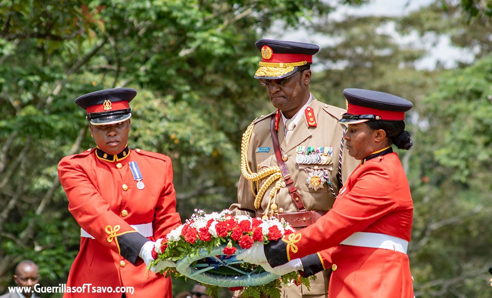 Members of the Kenyan Armed Forces holding a wreath on their way to lay it to a war grave. A general, complete with peaked black and gold cap and tan uniform with gold lanyard, stands behind two guards in bright red coats.