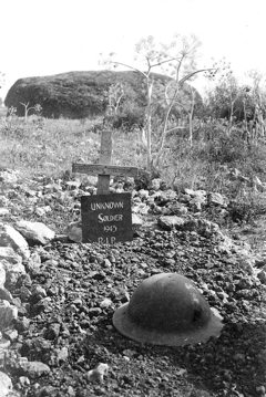 Wooden field grave marker for an unknown soldier killed in 1943, L'Esperance Farm, France. A Brodie helmet sits in front of the wooden cross atop a patch of freshly dug top soil, marking the soldier's grave