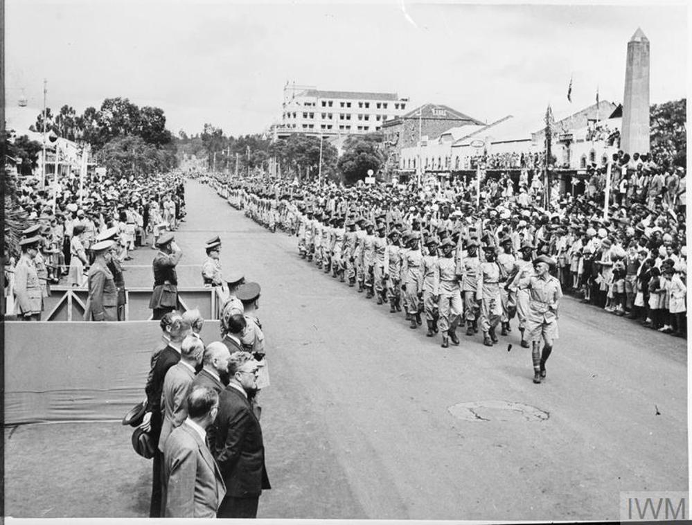 Members of the African Pioneer Corps parade through downtown Nairobi on VE Day 1945. They are flanked by crowds full of jubilant civilians, dignitaries, and military figures.