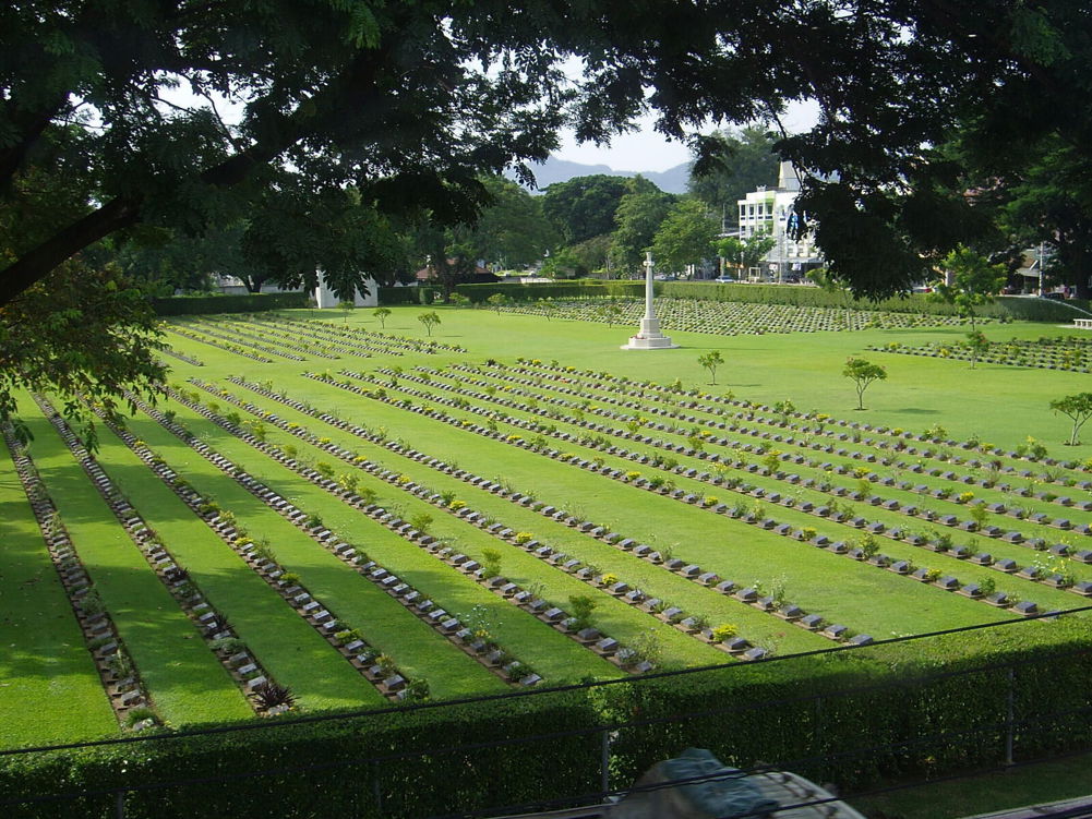 A wide view of the small black headstone plaques at Kanchanburi war cemetery. The POW grave markers stretch across the length of the cemetery. A white stone cross of sacrifice sits in the middle of the headstone rows. The graves themselves are set upon a crisp green lawn.