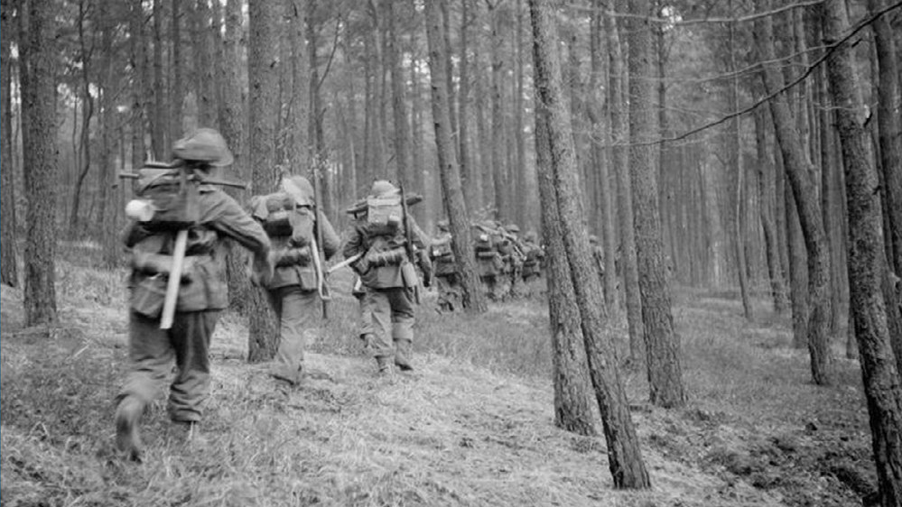 British infantry advances through the densley forested Reichswald Forest in Germany. The soldiers move in a single column one after another through a path amidst think tall tree trunks