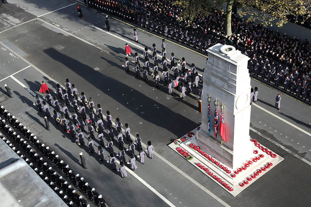 Soldiers in grey great coats parade past the Cenotaph on Remembrance Sunday. Many wreaths have been laid on the steps of the memorial, and several banners have been hung alongside it.
