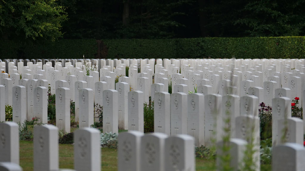 Rows of Commonwealth War Graves Commission headstones at Reichswald Forest War Cemetery. A large green boundary hedge is visible in the background with the lower branches of a green tree creeping into the top left corner.