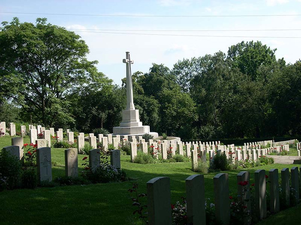The CWGC plot at Poznan Old Cemetery showing rows of white, round topped CWGC headstones with a Cross of Sacrifice among them. The headstone rows descend downhill to the right. A variety of lush green trees against a blue sky is visible in the background.
