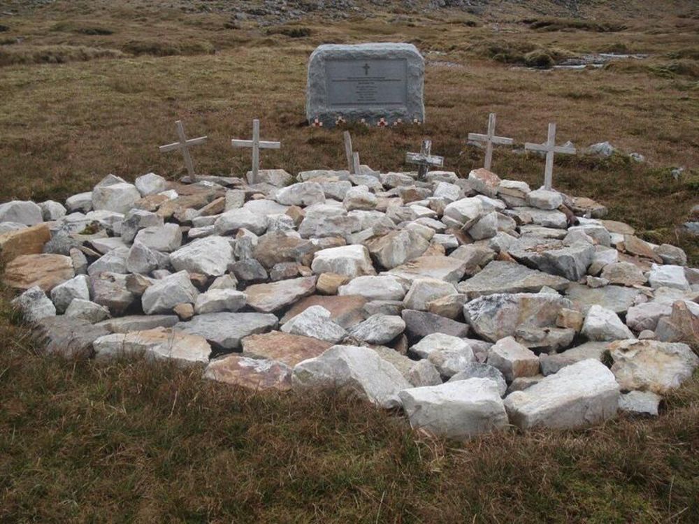 Stone cairn with six wooden cross-shaped grave markers. A large stone memorial to the people buried here is visible in the background.