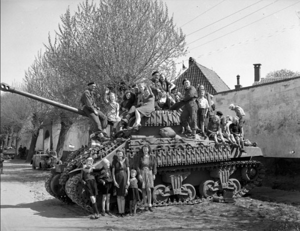 Dutch CHildren sitting on top of a Canadian Sherman Firefly tank in front of a high wall and a row of tall trees in the Netherlands, circa April 1945.