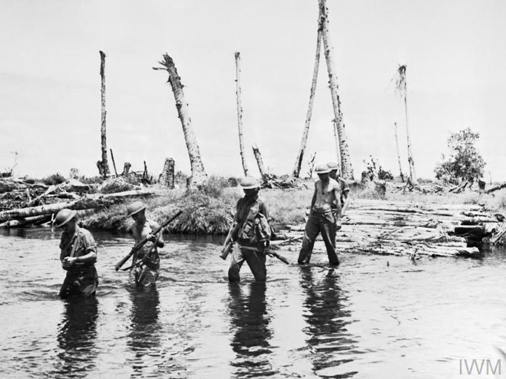 Australian soldiers wading through a deep river after stepping off a rickety wood-slatted wharf in northern New Guinea. The river bed shows dead, damaged trees, probably the result of artillery fire.