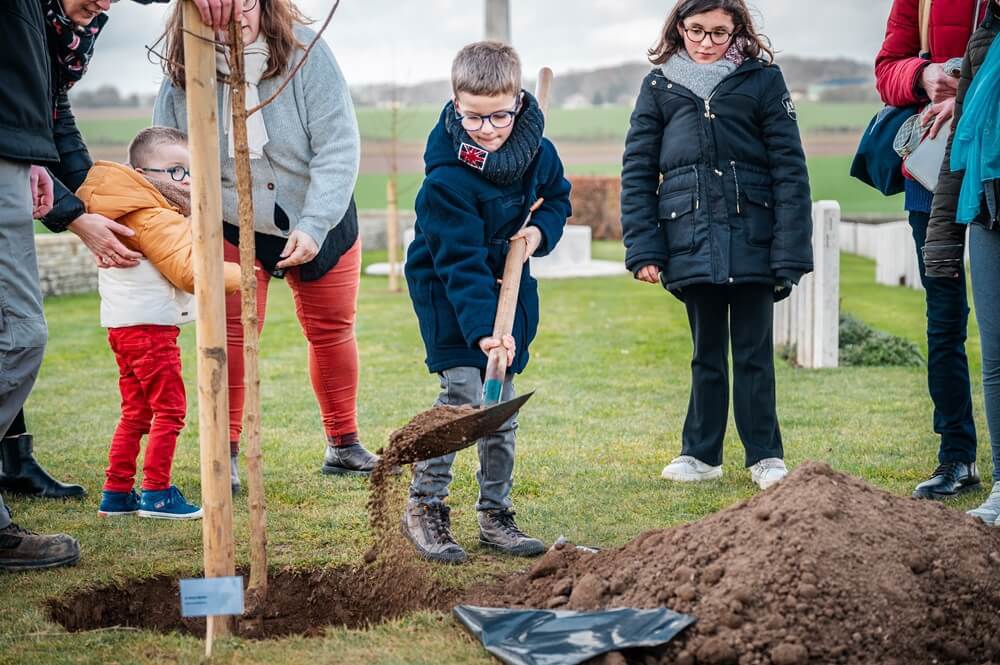 Young child in glasses shovels soil into the base of a fresh sapling. His friends and family are watching in the background.