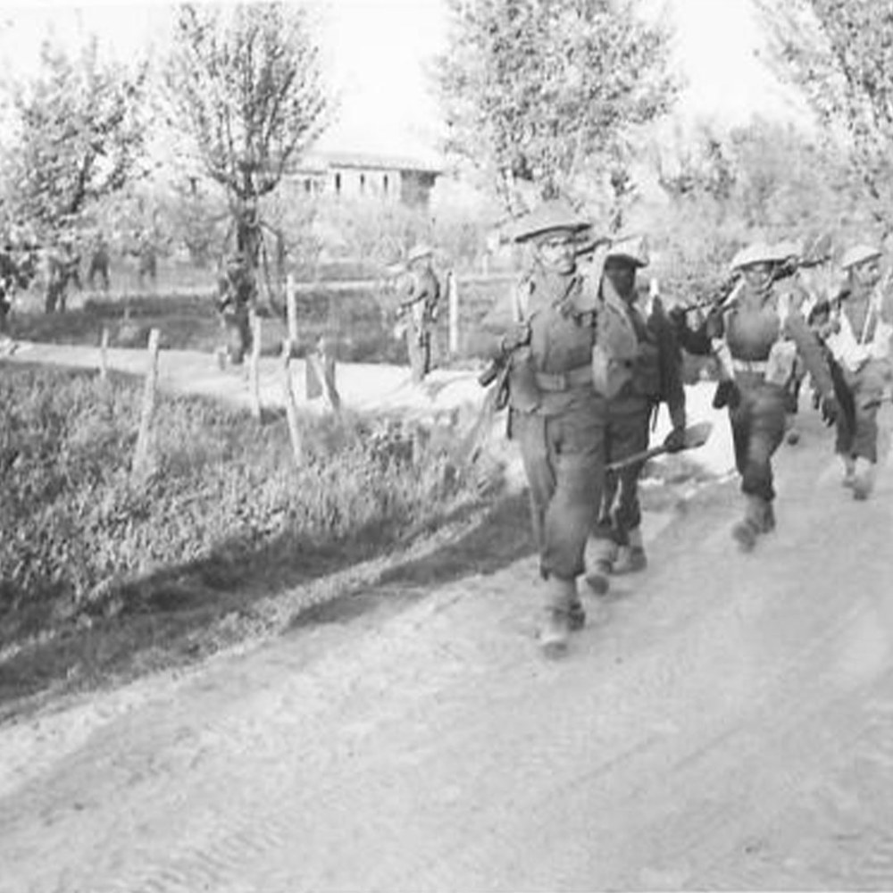 Men of 1st/5th Mahrattas arrive at start point for attack in Italy, circa early 1945. A low farm building is visible in the background. The road they are advancing on is flanked by trees on one side and a low wire fence on the other. © IWM