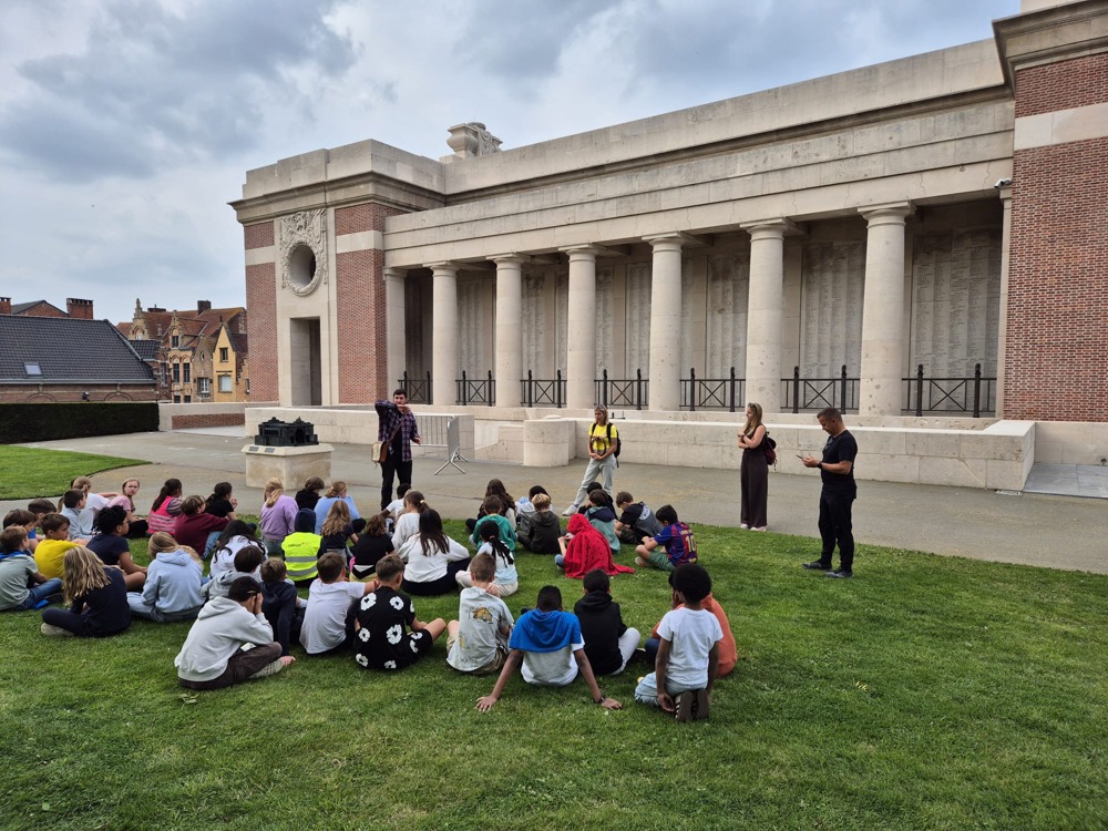 Tour guides speaking to a group of seated adolecents on the Rampart side of the Menin Gate Memorial, Ypres