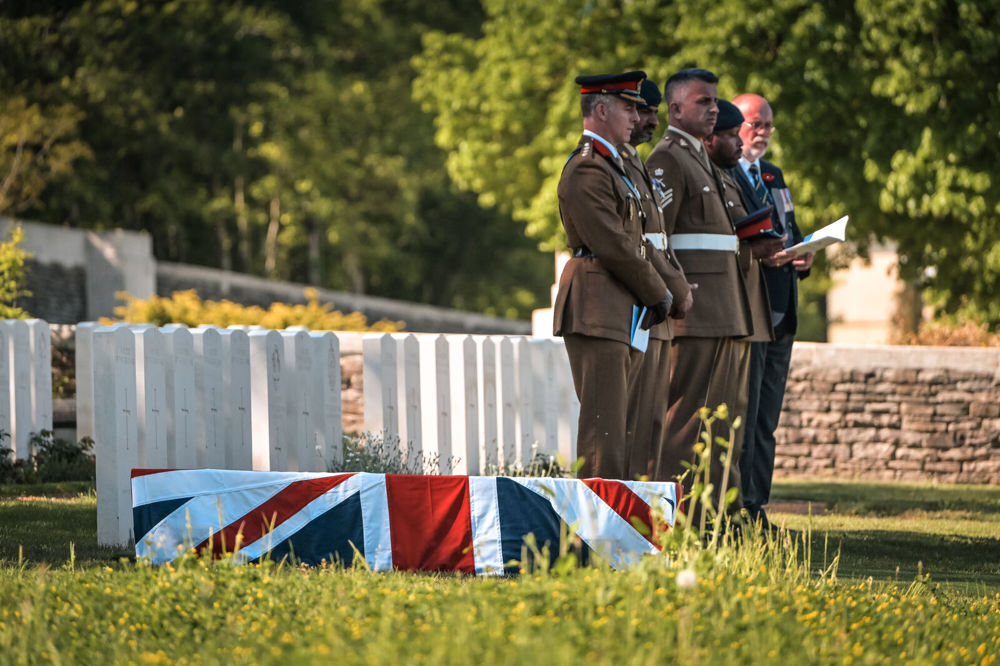A Union Jack drapped coffin lies on the floor waiting for internment next to a row of CWGC headstones. A group of soldiers waits patiently next to the coffin.