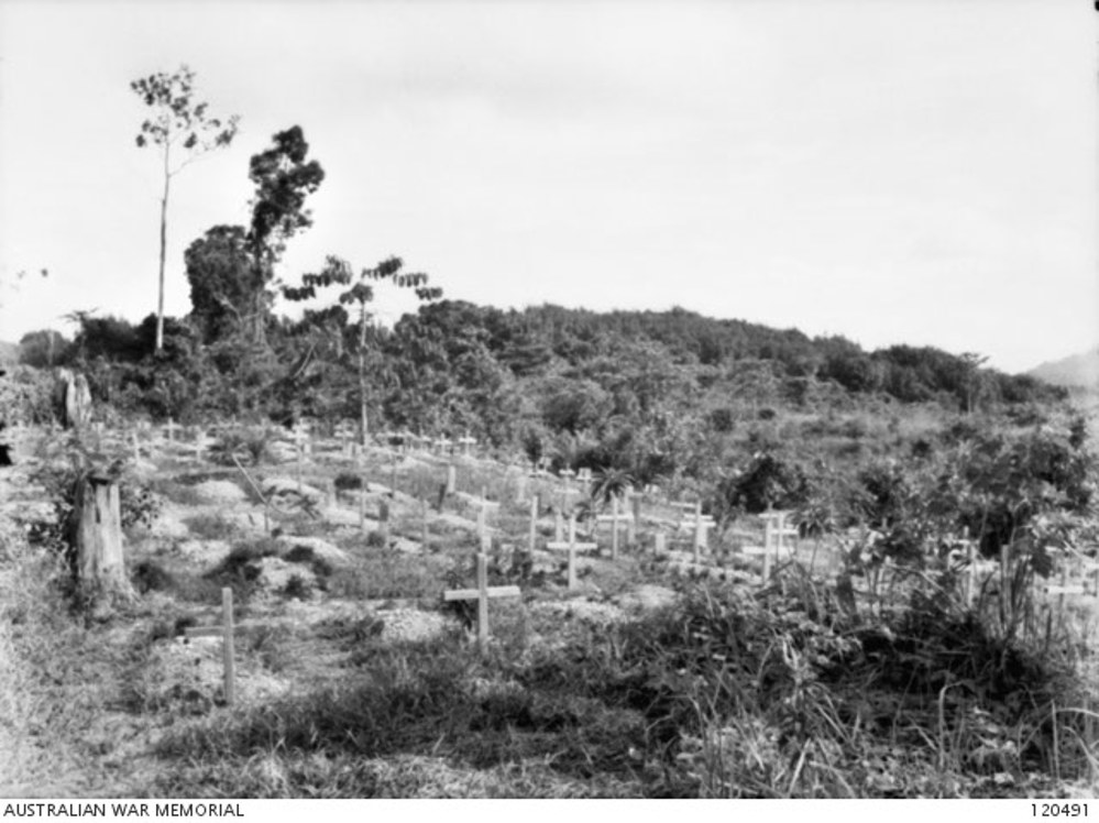 Scene of a rudimentary cemetery made for service men on a bare hillside in Sandakan, Indonesia following the Sandakan Death March. The graves of various POWs are visible, marked with simple wooden crosses. In the background are jungle covered hills.