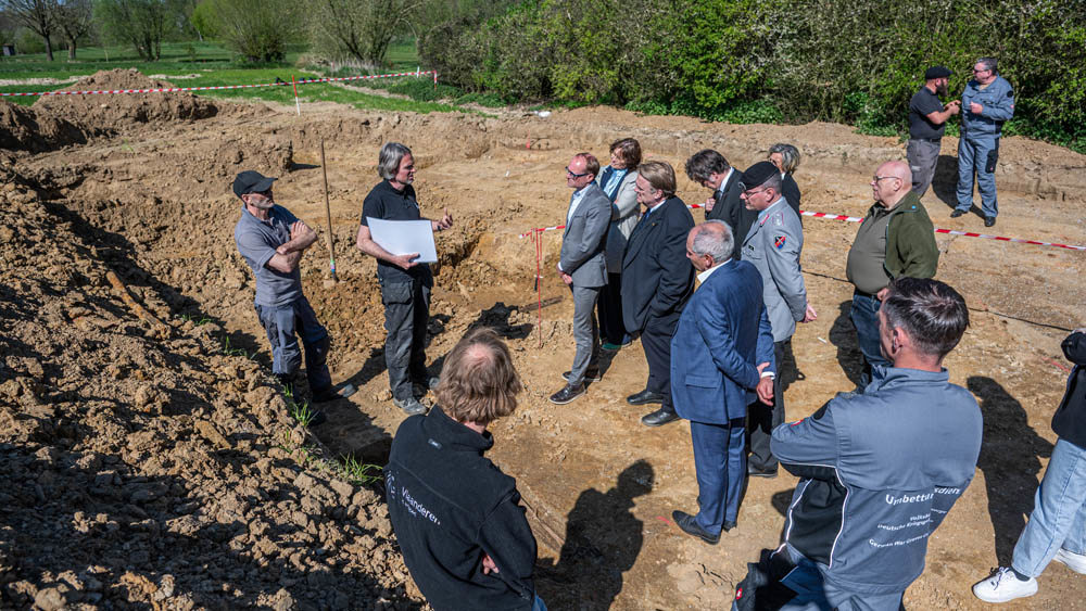 A group of men, some in work clothes, some in suits, in a discussion next to a large archaeological trench.