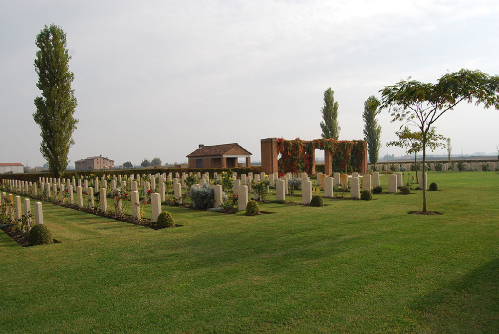 View of Argenta Gap War Cemetery showing rows of headstones, tall trees, the red brick shelter, and the central