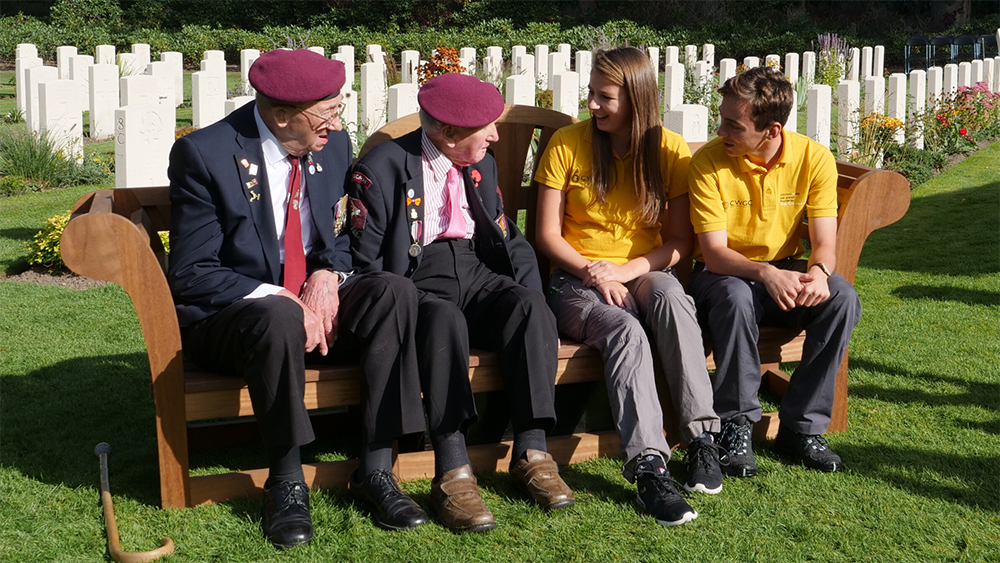 WW2 Veterans chat with CWGF guides on a bench in front of rows of CWGC headstones.